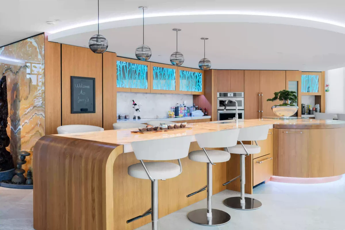 Modern kitchen with blonde wood cabinets, a large island with three white bar stools, and blue backlit accent lighting.