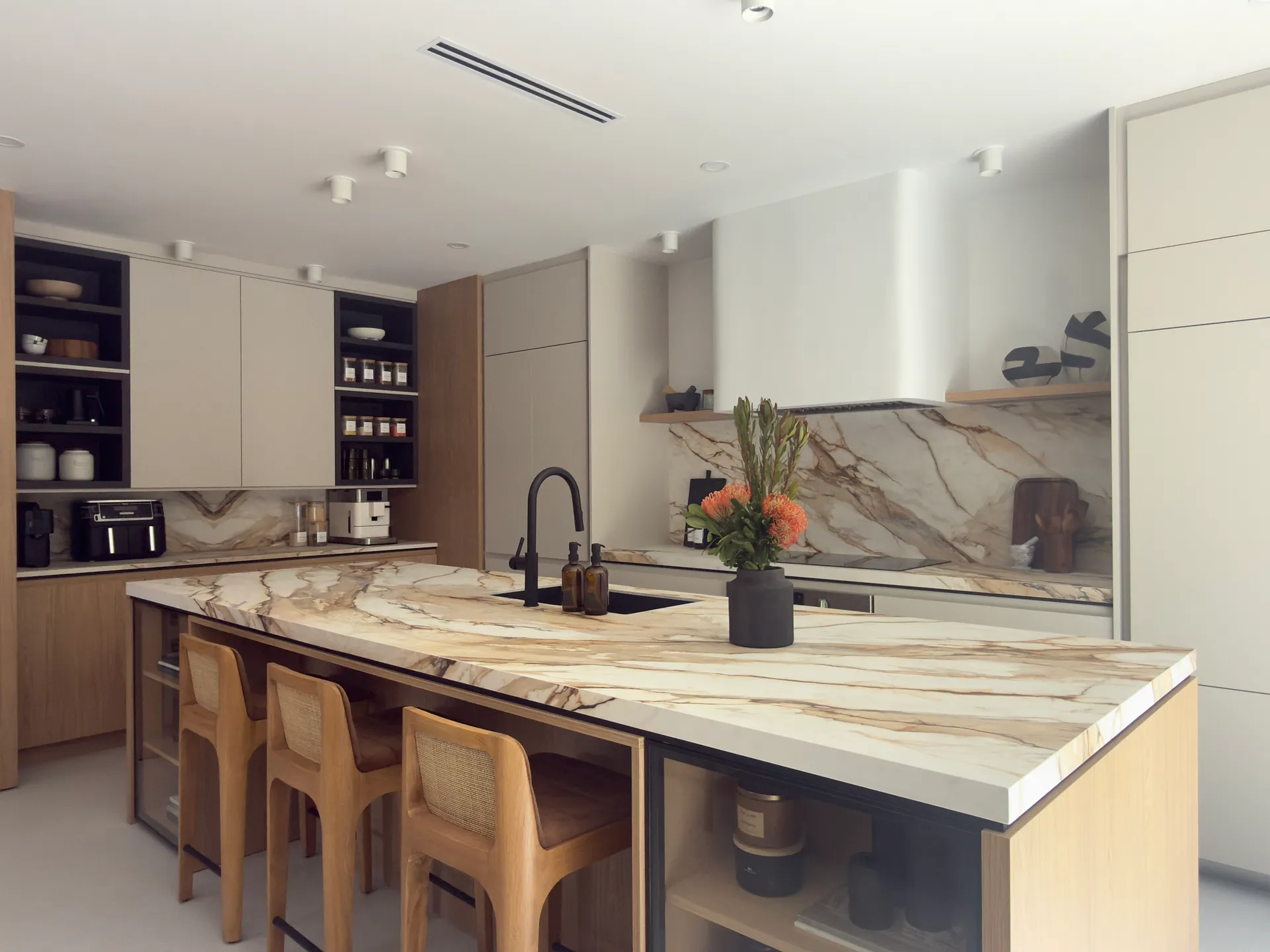 Modern kitchen with a large marble island countertop, sink, black faucet, three wooden bar stools, and grey/wood cabinets.