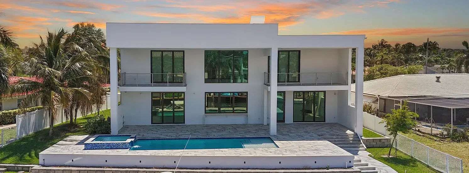 Modern white two-story house with black-framed windows, a pool, and hot tub under an orange sunset sky.