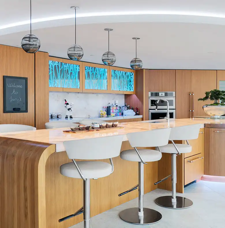 Modern kitchen with a curved wooden island, white bar stools, striped spherical pendant lights, and blue-lit cabinetry.