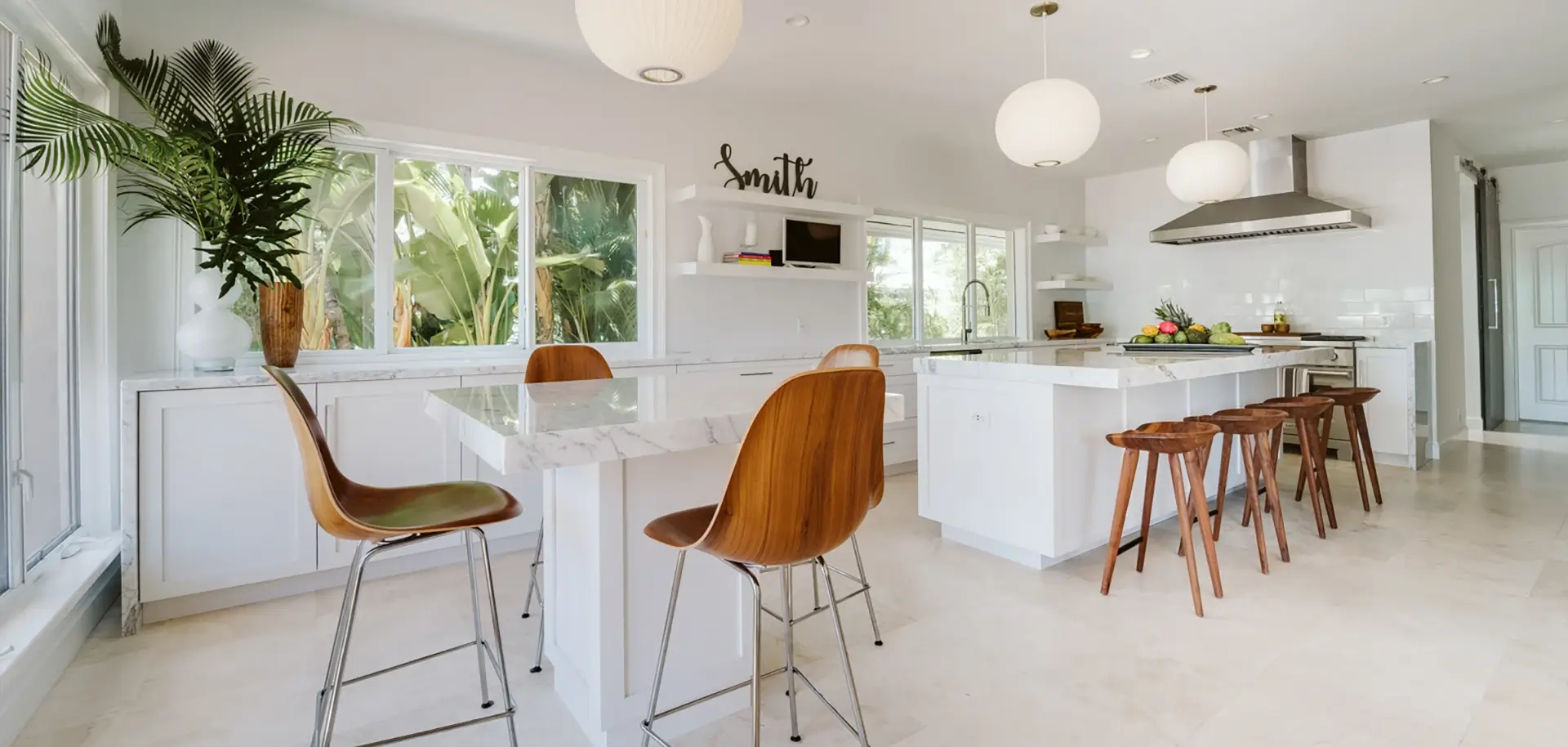 Bright, modern kitchen with white cabinets, marble countertops, multiple barstools, and large windows revealing lush tropical plants.
