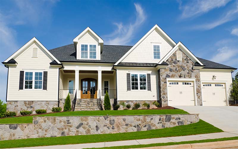 Large cream house with dark roof, stone accents, front porch with columns, and two garage doors, on a green lawn.