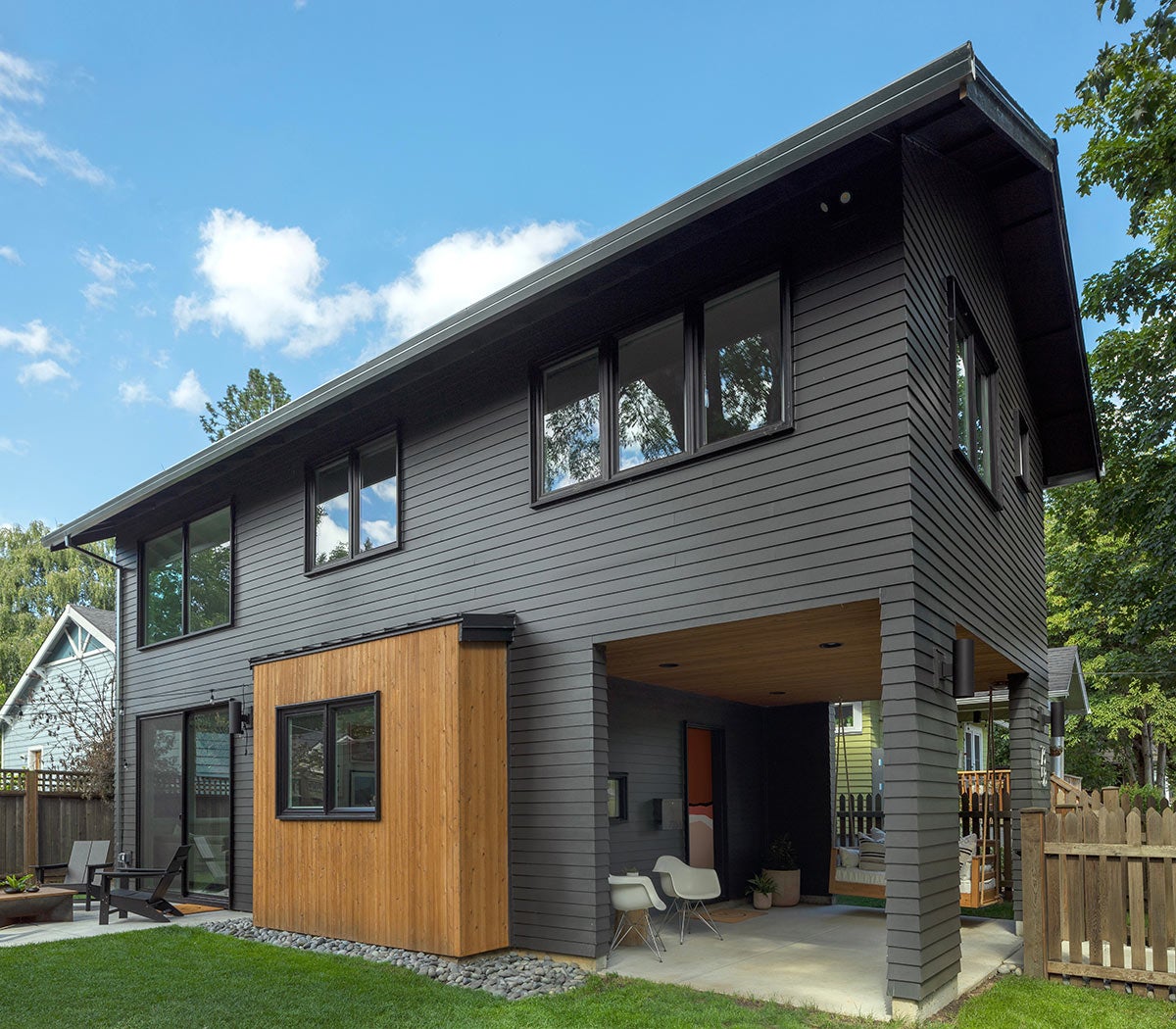 Modern two-story house with dark gray siding, wood accents, a grassy lawn, and covered outdoor patio areas.