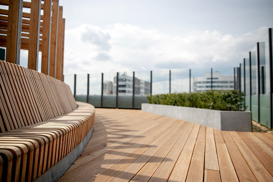 Curved wooden bench and deck on a modern rooftop terrace with glass barriers and city views.