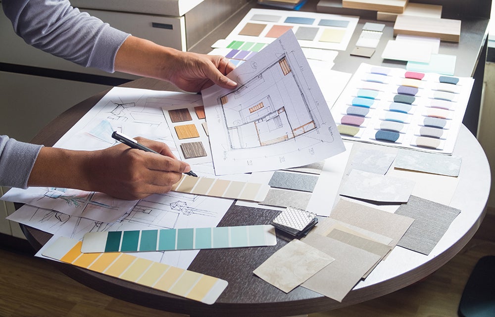 Designer's hands working on architectural plans with color swatches and material samples on a table.