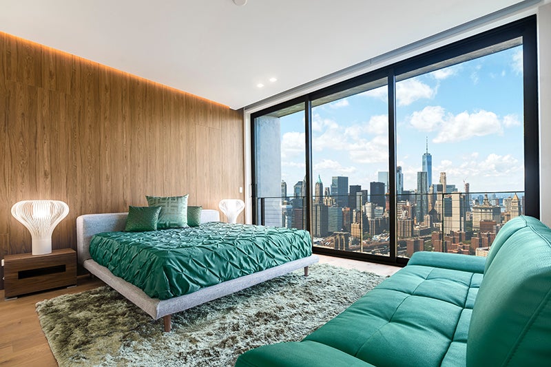 Modern bedroom with wood-paneled wall, green bed, furry rug, and a large window overlooking a city skyline.