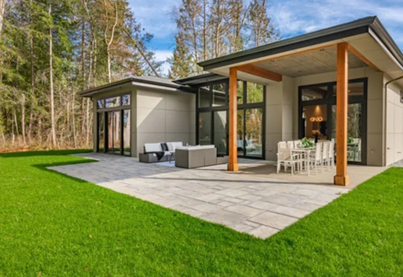 Modern house with a large patio featuring outdoor dining and lounge areas, green lawn, and pine forest backdrop.