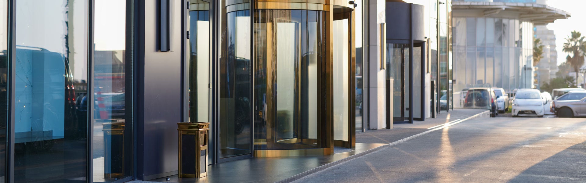 Modern building entrance with a prominent brass and glass revolving door, reflections, and a parking lot with cars.