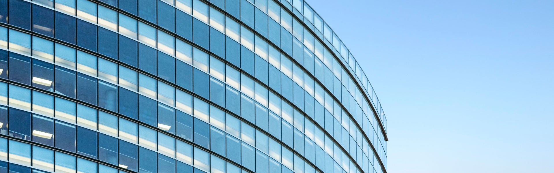Curved glass building facade with numerous windows reflecting light against a blue sky.
