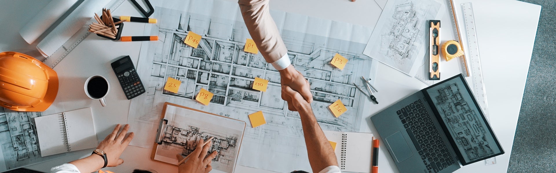 Two people shake hands over architectural blueprints and tools on a desk, seen from above.