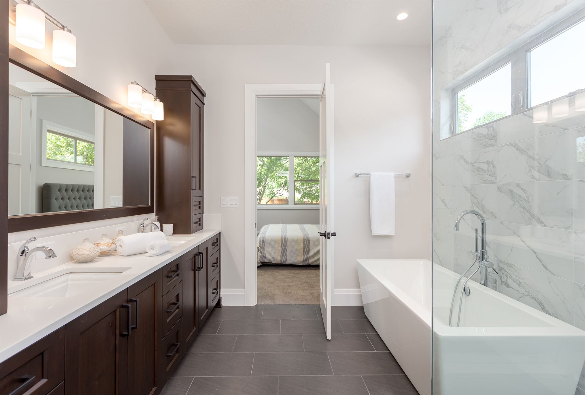 Modern bathroom with dark wood double vanity, freestanding tub, and a bedroom visible through an open door.