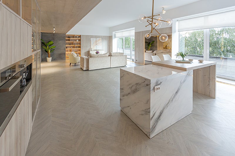 Modern open-plan kitchen and living room with marble island, herringbone wood floor, and large windows.