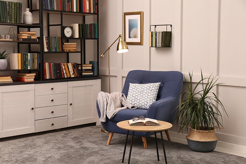 A cozy reading nook with a blue armchair, a golden floor lamp, a bookshelf, and a small table with an open book.