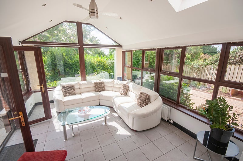 Bright sunroom with a white sectional sofa, glass coffee table, tiled floor, and large windows overlooking a green garden.
