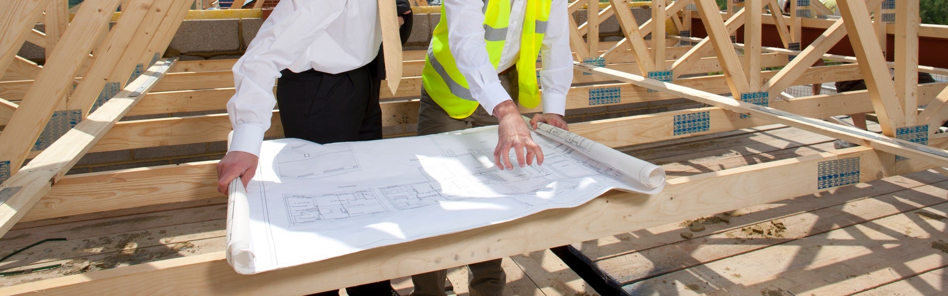 Two people reviewing blueprints laid on wooden roof trusses at a construction site.