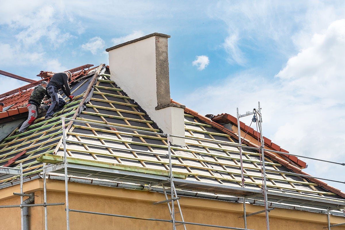 Two workers on a roof under repair with exposed wooden slats, black underlayment, and a chimney, with scaffolding.